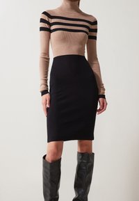 Donna che indossa un vestito a coste beige e nero, aderente, abbinato a stivali in pelle nera fino al ginocchio, su uno sfondo semplice.