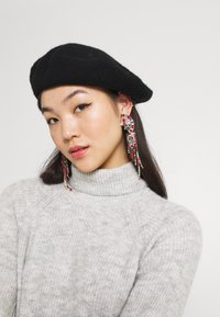 Juodas veltinis beretė su minkšta tekstūra, derinama su ilgomis karoliukų auskarais, turinčiais spalvingų dekoracijų, dėvima su šviesiai pilku megztu turtleneck svarku.