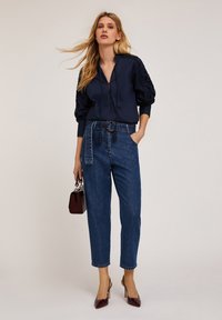 Marinblus med puffärm och spetsdetaljer, tillsammans med högmidjade ljusblå jeans och bordeauxfärgade stilettklackar, håller en mörk handväska.