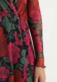 Langarmkleid aus einem schwarzen durchsichtigen Stoff mit leuchtend pinken und roten Blumenmustern, detaillierten grünen Blättern und einem gerafften Bund.