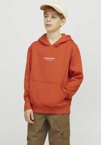 Felpa arancione con tasca a marsupio e testo bianco che recita "Originals Studio". Abbinata a un cappellino beige e pantaloni cargo verdi oliva.