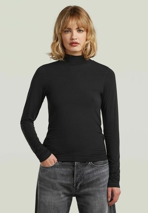 Kobieta z blond włosami do ramion, ubrana w czarny, długi sweter z golfem i szare dżinsy, stoi z jedną ręką w kieszeni.