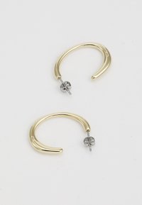 Orecchini a cerchio in metallo color oro con un design morbido e curvo. Presentano un perno in metallo color argento e una chiusura sul retro. Notabili per il loro stile minimalista.