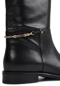 Bota de cuero negro hasta la rodilla con un diseño elegante, acento de cadena dorada y costuras minimalistas sobre una suela plana negra.