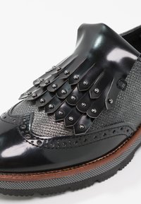Mocassin en cuir noir et tissu texturé avec franges décoratives et clous en métal, détails brogue, et semelle superposée avec coutures.