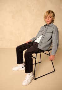 Veste grise boutonnée, t-shirt blanc, pantalon en velours côtelé foncé, baskets blanches, assis sur une chaise en métal noir sur un fond beige.