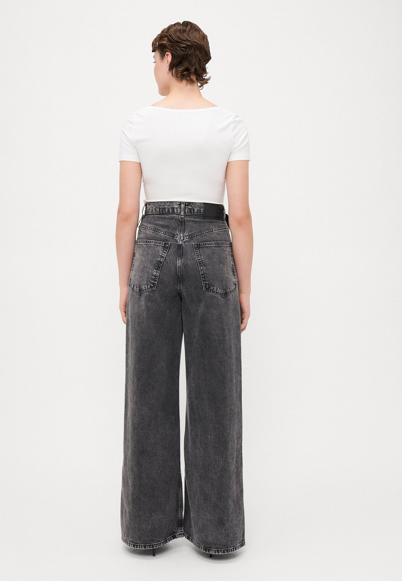 Jeans a gamba larga realizzati in denim grigio scuro con vita alta, dotati di tasche posteriori e orlo cucito. Abbinati a una maglietta bianca a maniche corte.