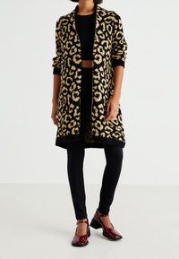 Leopardmönstrad lång kappa i svart och beige stickad, med öppen front och ribbade ärmslut, tillsammans med svarta leggings och vinröda klackskor.