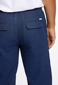 Mörkblå texturerade byxor med två bakfickor och en liten etikett. Tyget verkar mjukt och något texturerat.