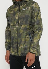 Man in groene camouflage Nike-jack met het tekstpatroon "Just Do It", zwarte broek, staand met ontspannen handen langs zijn zij.