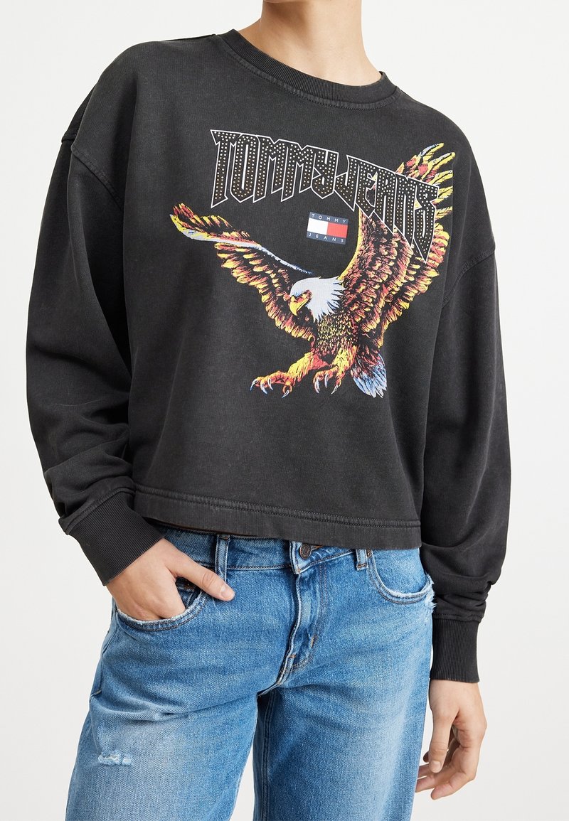 Zwarte cropped sweatshirt met een afbeelding van een arend in levendige kleuren en de tekst "TOMMY JEANS" erboven. Lange mouwen met ribgebreide manchetten.