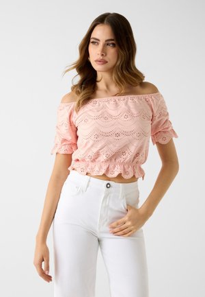 Frau trägt ein pinkfarbenes, schulterfreies Lochmuster-Crop-Top und hoch taillierte weiße Hose, steht vor schlichtem, hellem Hintergrund.