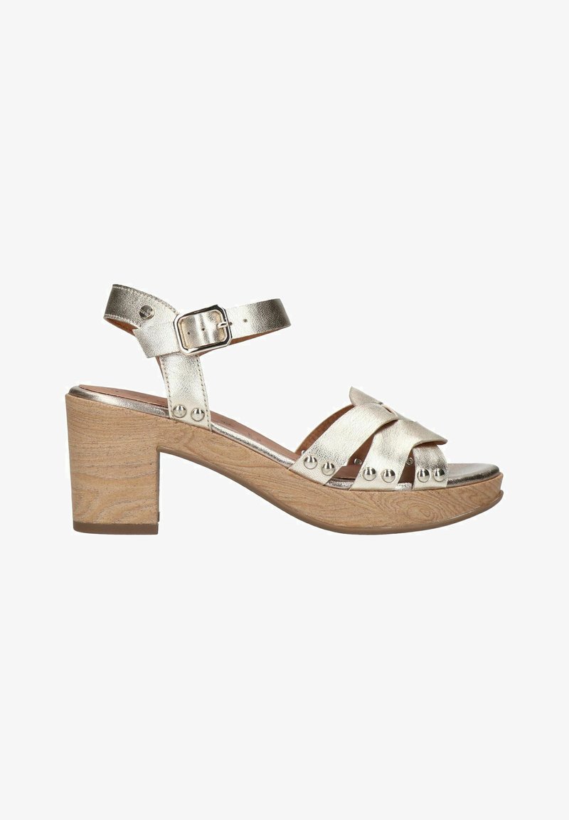 Silver metallic heeled sandals with a wooden block heel, featuring crossed straps and metal studs. Adjustable ankle strap for fit.