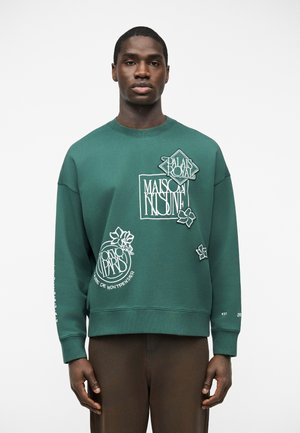 Homme portant un sweat-shirt vert avec un texte graphique blanc et des motifs floraux, associé à un pantalon marron, debout devant un fond blanc.