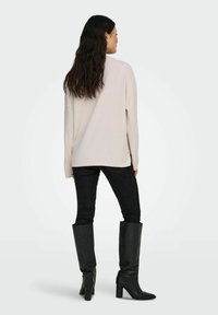 Suéter de punto beige claro con hombros caídos, combinado con jeans negros ajustados y botas negras de tacón alto hasta la rodilla. Diseño simple, textura suave.