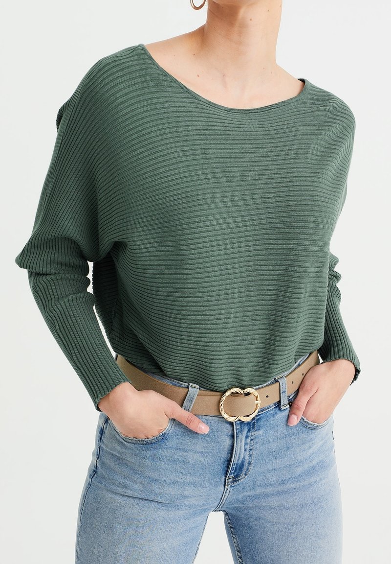 Femme portant un pull côtelé vert rentré dans un jean bleu clair avec une ceinture beige avec une boucle circulaire dorée, mains dans les poches.