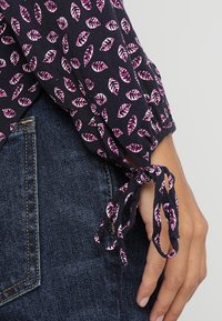 Manica in tessuto navy con motivo a foglia rosa, legata a fiocco sopra il polso, accanto a una mano appoggiata sulla tasca di jeans denim blu scuro.