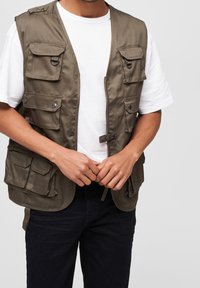 Olivgrüne Utility-Weste mit mehreren Taschen, Druckknopfverschlüssen und Stoffstruktur. Wird über einem weißen T-Shirt getragen und mit dunklen Jeans kombiniert.