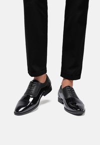 Scarpe da dress nere con una finitura in vernice lucida e una parte superiore in rete testurizzata, caratterizzate da un design con lacci e punta arrotondata.