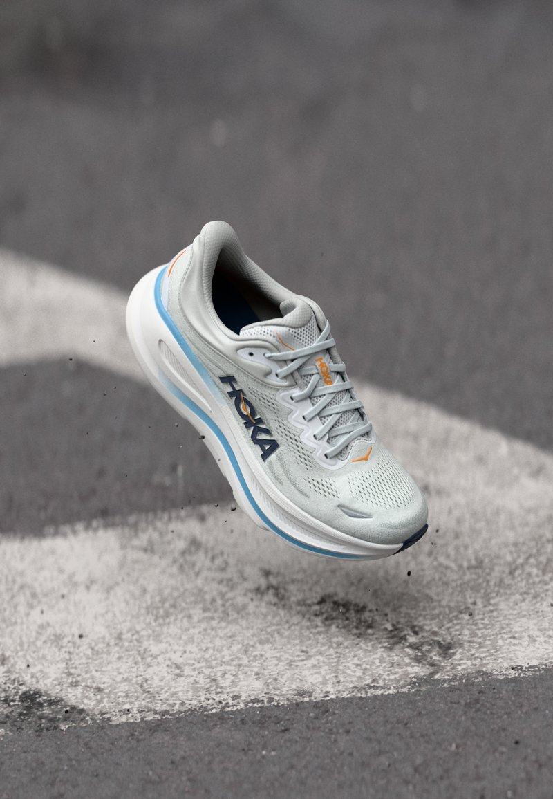 Sapatilha de corrida cinza claro com parte superior em malha respirável, detalhes em laranja e uma entressola branca grossa com um acabamento azul. Design leve.