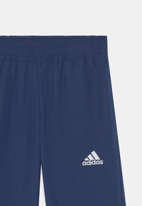 Tamsiai mėlynos sportinės kelnės su elastine juosta ir subtilia tekstūra. Dešinėje šlaunyje yra balta Adidas logo.