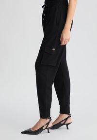 Pantaloni da jogging neri con vita elasticizzata, tasche laterali e una grande tasca cargo sulla coscia, abbinati a décolleté nere con cinturino alla caviglia.