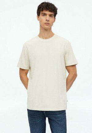Junger Mann mit lockigem Haar, der ein schlichtes beiges kurzärmeliges T-Shirt und blaue Jeans trägt, stehend mit den Händen hinter dem Rücken.