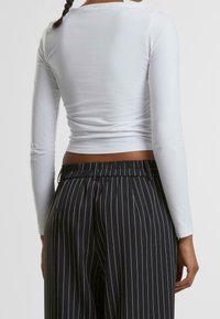 Weißes Langarmoberteil mit körperbetontem Schnitt, kombiniert mit hochtaillierten, schwarz gestreiften Hosen, die über einen elastischen Bund und Faltendetails verfügen.