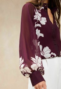 Burgundy blouse in sheer fabric with floral white print, high collar, button-down front, and puffed long sleeves with fitted cuffs.