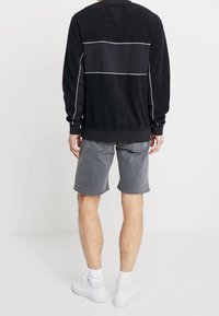 Persona in piedi di spalle, indossa una maglietta nera a maniche lunghe con strisce bianche, pantaloni corti di jeans grigi, calzini bianchi e sneaker bianche.