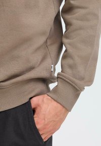 Brun genser i myk stoff med ribbestrikkede mansjetter og nederkant, har en side søm etikett og avslappet passform. Hånd som hviler i en lomme.