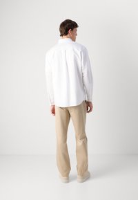 Man die met zijn gezicht weg van de camera staat, gekleed in een witte overhemd met knopen, beige broeken en beige sneakers, tegen een effen witte achtergrond.