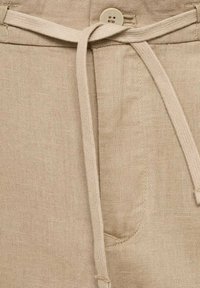 Shorts en lin beige avec un lien à nouer, fermeture à bouton et une poche latérale. Le tissu a une apparence lisse et texturée.