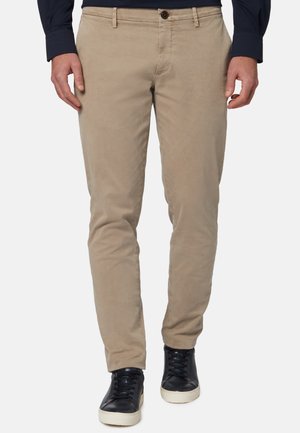 Beige Chinos aus Baumwolle mit glatter Textur, schlanker Schnitt, Fronttaschen und Ein-Knopf-Verschluss. Kombiniert mit schwarzen Sneakers.