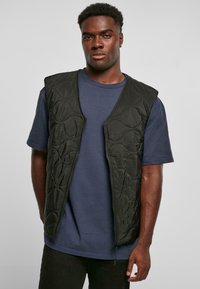 Zwarte gewatteerde vest met een rits aan de voorkant, met een gestructureerd oppervlak en zonder mouwen, gedragen over een blauw T-shirt met korte mouwen.
