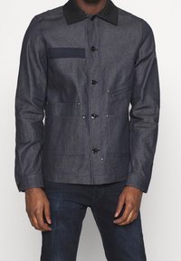 Denim jacket in dark blue with a black collar, featuring a chest pocket, two front pockets, and button closure. Smooth texture, tailored fit.