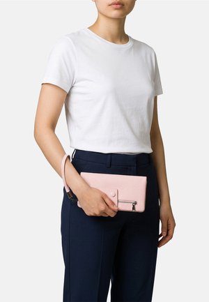Donna con una maglietta bianca a maniche corte e pantaloni blu navy che tiene una pochette da polso rosa chiaro con trama e tasca con cerniera.