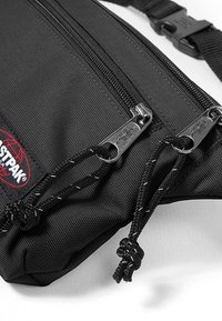 Mustakangas ristiolkalaukku kahdella vetoketjullisella osastolla, jossa on teksturoitu pinta, säädettävä hihna ja brändilogo.