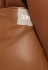 Brun lærveske med en teksturert overflate. Har en grå etikett som viser "MM" i hvit tekst, delvis dekket av et brunt stoff.