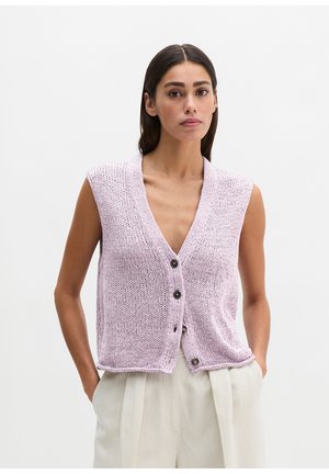 Kobieta z prostymi ciemnymi włosami, ubrana w bezrękawnik z dzianiny w kolorze lawendowym, zapinany na guziki, oraz jasnobeżowe plisowane spodnie, z rękami w kieszeniach.
