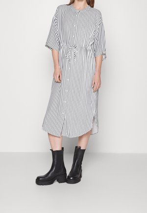 Kvinde iført en hvid midilang kjole med lodrette sorte striber og sorte chunky ankelstøvler, stående mod en ensfarvet hvid baggrund.