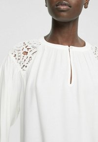 Weiße Bluse mit rundem Ausschnitt, ausgestattet mit einer Teilknopfleiste und Spitzenverzierungen an den Schultern. Weicher, fließender Stoff mit Falten.