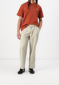 Camiseta de algodón color óxido con mangas cortas, combinada con pantalones plisados de color beige claro. Zapatillas negras con franjas blancas completan el conjunto.