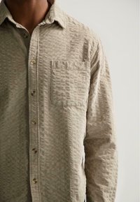 Primer plano de una persona que lleva una camisa de botones de manga larga texturizada color beige con un bolsillo en el pecho, frente a un fondo claro y liso.