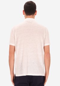 Un uomo di spalle indossa una polo bianca trasparente a maniche corte e pantaloni scuri su uno sfondo chiaro e semplice.