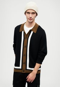 Fekete kardigán barna gallérral és fehér csíkokkal, gombos elülső dizájnnal, textúrált kötött anyagból készült, hosszú ujjal. Bézs sapka van rajta.