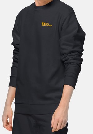 Osoba nosząca czarną bluzę z długim rękawem z żółtym logo Jack Wolfskin na lewej piersi na jednolitym tle.