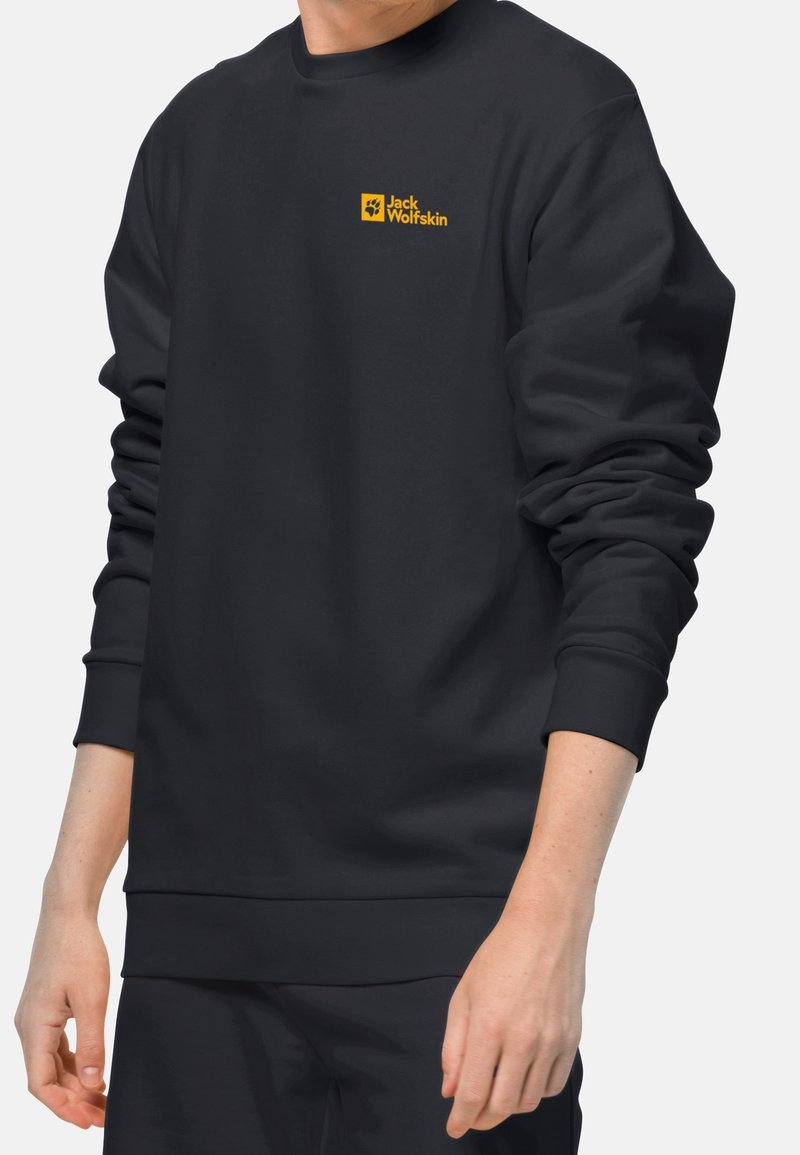 Personne portant un sweat-shirt noir à manches longues avec un logo jaune Jack Wolfskin sur la poitrine gauche sur un fond uni.