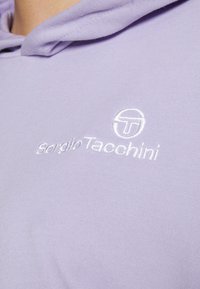 Sudadera de color púrpura claro hecha de tejido suave. Presenta un texto de logo blanco bordado "Sergio Tacchini" y un diseño de logo circular.