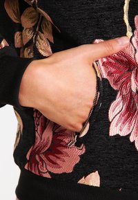 Černá mikina s výraznou květinovou výšivkou v růžové a vínové barvě, s kapsou na zip na boku a žebrovanými manžetami.